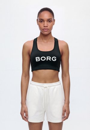 Une femme porte un soutien-gorge de sport BORG noir et un short blanc à cordon sur un fond clair et uni.