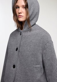 Cappotto di lana grigio con cappuccio, due grandi bottoni frontali e una tasca laterale con una finitura morbida e testurizzata.