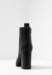 Musta nahkainen nilkkuri, jossa on jykevä lohkokorko, sileä pinta, pyöristetty varvas ja takana vetoketjuluukku. Sivussa näkyvää vetoketjydetailia.