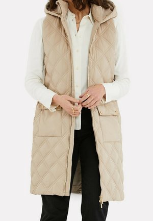 Femme portant un gilet matelassé beige sans manches à capuche par-dessus une chemise blanche boutonnée et un pantalon noir, debout les mains jointes près de la taille.
