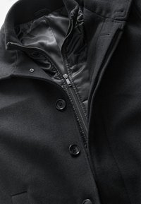 Zwart wollen mantel met een gewatteerde voering, voorzien van een ritssluiting, knoopdetails en een gestructureerde kraag. Overal een gladde textuur.