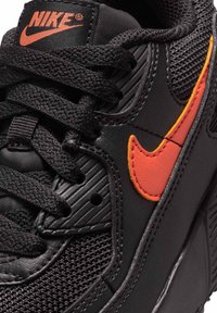 Close-up van een zwarte Nike-sneaker met gestructureerde veters en oranje swoosh-logo aan de zijkant en op de tong.