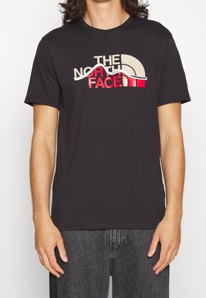 Osoba nosi czarną koszulkę z krótkim rękawem z logo The North Face w beżowo-czerwonych kolorach na piersi, zestawioną z ciemnoszarymi dżinsami.