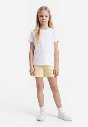 Blondynka stojąca, ubrana w prostą białą koszulkę, jasnżółte szorty, białe skarpetki i białe tenisówki na białym tle.