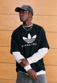 Svart oversized t-shirt med vit text "BASKETBALL" och logotyp, lager-på-lager över en vit långärmad tröja. Grå keps och texturerat kedjehalsband.