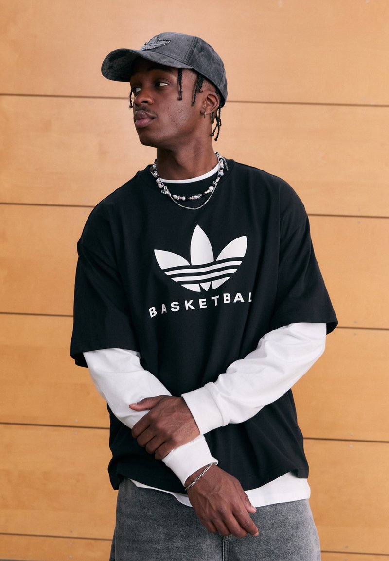 Svart oversized t-shirt med vit text "BASKETBALL" och logotyp, lager-på-lager över en vit långärmad tröja. Grå keps och texturerat kedjehalsband.