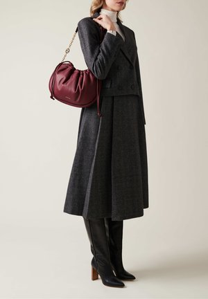Costume en mélange de laine gris avec un manteau croisé, jupe plissée, bottes noires en cuir à talons hauts arrivant au genou et un sac en cuir bordeaux à cordon.