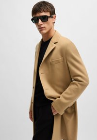 Cappotto in lana beige con vestibilità sartoriale, tasca sul petto singola e chiusura con bottoni. Indossato sopra un maglione nero, abbinato a occhiali da sole neri a forma quadrata.