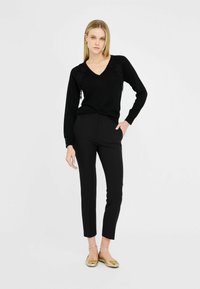 Schwarzer V-Ausschnitt-Pullover mit Spitze an den Schultern, kombiniert mit maßgeschneiderten schwarzen Hosen und goldenen Ballerinas. Glatter Stoff, figurschnitt.