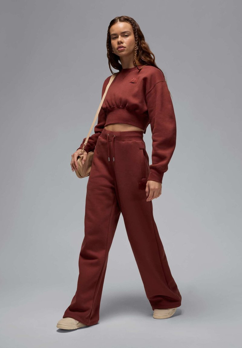 Bordeauxroter, kurzer Sweatshirt mit langen Ärmeln, kombiniert mit passenden, lässigen Jogginghosen. Das Modell hält eine kleine Handtasche; subtile Texturen sind sichtbar.