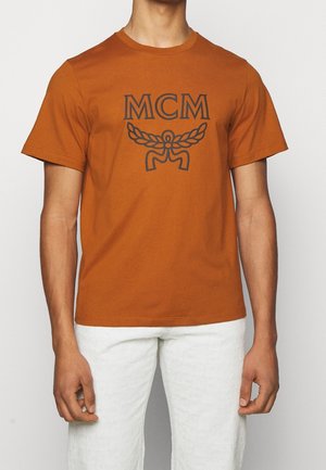 Man draagt een roestkleurig MCM-logo T-shirt en gestructureerde witte broek, staand tegen een effen lichte achtergrond.