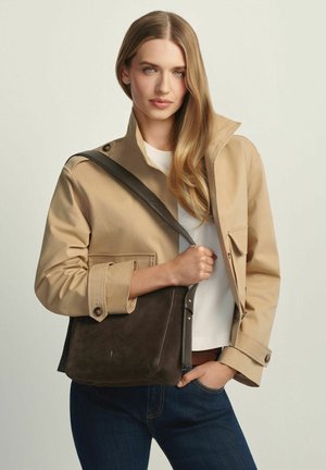 Blondynka w beżowej kurtce i białym topie niesie ciemnobrązową skórzaną torbę na ramię, stojąc na tle jednolitego jasnego tła.