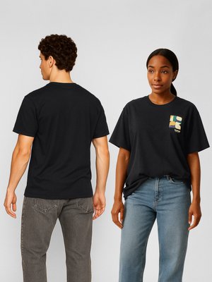 Mand, der viser ryggen, og kvinde, der vender fronten, iført sorte grafiske t-shirts og afslappede jeans mod en ensfarvet baggrund.