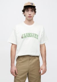 Λευκό βαμβακερό μπλουζάκι με πράσινο floral κείμενο "CARHARTT." Φορεμένο με χακί παντελόνι. Το μοντέλο έχει κοντά, σγουρά μαλλιά και μπεζ καπέλο.