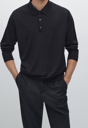 Mann trägt ein schwarzes langärmeliges Polohemd mit drei Knöpfen und schwarze Hose, Hände in den Hosentaschen.