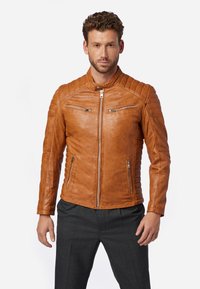 Tan leren jack met gequiltte schouders en ritszakken. Heeft een staande kraag en een gladde textuur. Voorzien van een ritssluiting aan de voorkant.