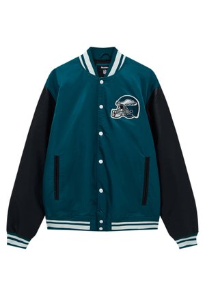 Personne portant une veste Eagles NFL verte et noire, un pantalon noir, des baskets blanches et une chemise blanche à rayures verticales, debout dos à nous.