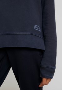 Sweat-shirt et pantalon bleu marine, gros plan sur le bas du torse et le bras droit avec un petit logo « Marc O » près de l’ourlet.