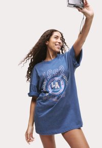 Blue oversized t-shirt with short sleeves, featuring a circular graphic design with "1992," "LA," and "WORLD TOUR" text in decorative font.