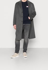 Grijze wollen jas met een getailleerd ontwerp, zwarte knopen, gedragen over een marineblauwe sweater, gecombineerd met grijze jeans en lichtgekleurde sneakers.