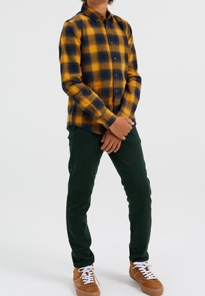 Niño de pie con las manos entrelazadas, vistiendo una camisa de cuadros amarilla y azul marino, pantalones verde oscuro y zapatillas marrones con cordones blancos.