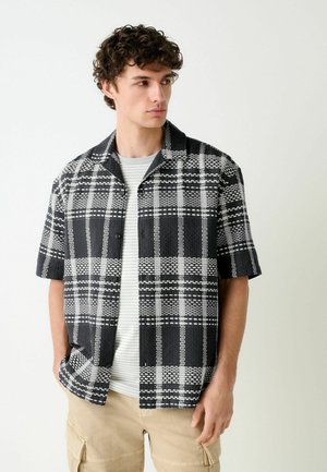 Junger Mann mit lockigem Haar, der ein schwarzes und weiß kariertes kurzärmeliges Hemd über einem hell gestreiften T-Shirt und beige Cargohosen trägt.