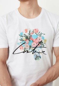 Valkoinen puuvillainen t-paita, jossa on värikäs kukkakuvio ja sana "Couture" lihavoidulla mustalla kursive-kirjaimilla. Lyhyet hihat ja pyöreä pääntie.