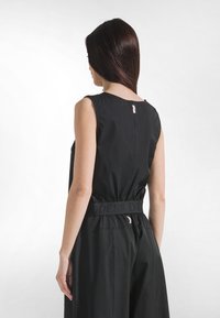 Schwarzer Jumpsuit mit tailliertem Bund und weitem Bein, mit rundem Ausschnitt und Schulterriemen. Mit einem strukturierten Taillenbanddetail.