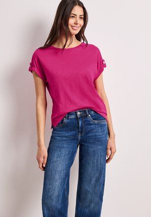 Kvinde står smilende iført en knaldrød kortærmet top med knappede opkørte manchetter og blå højtaljede viddebukser.