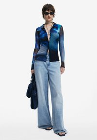 Long-sleeve mesh shirt with a blue abstract pattern, paired with light blue wide-leg jeans and a dark blue handbag. Black sunglasses accent the look.