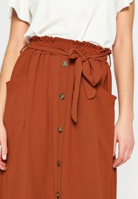 Gonna color ruggine con finitura testurizzata, vita elasticizzata, cintura annodata e chiusura con bottoni. Presenta due tasche laterali.