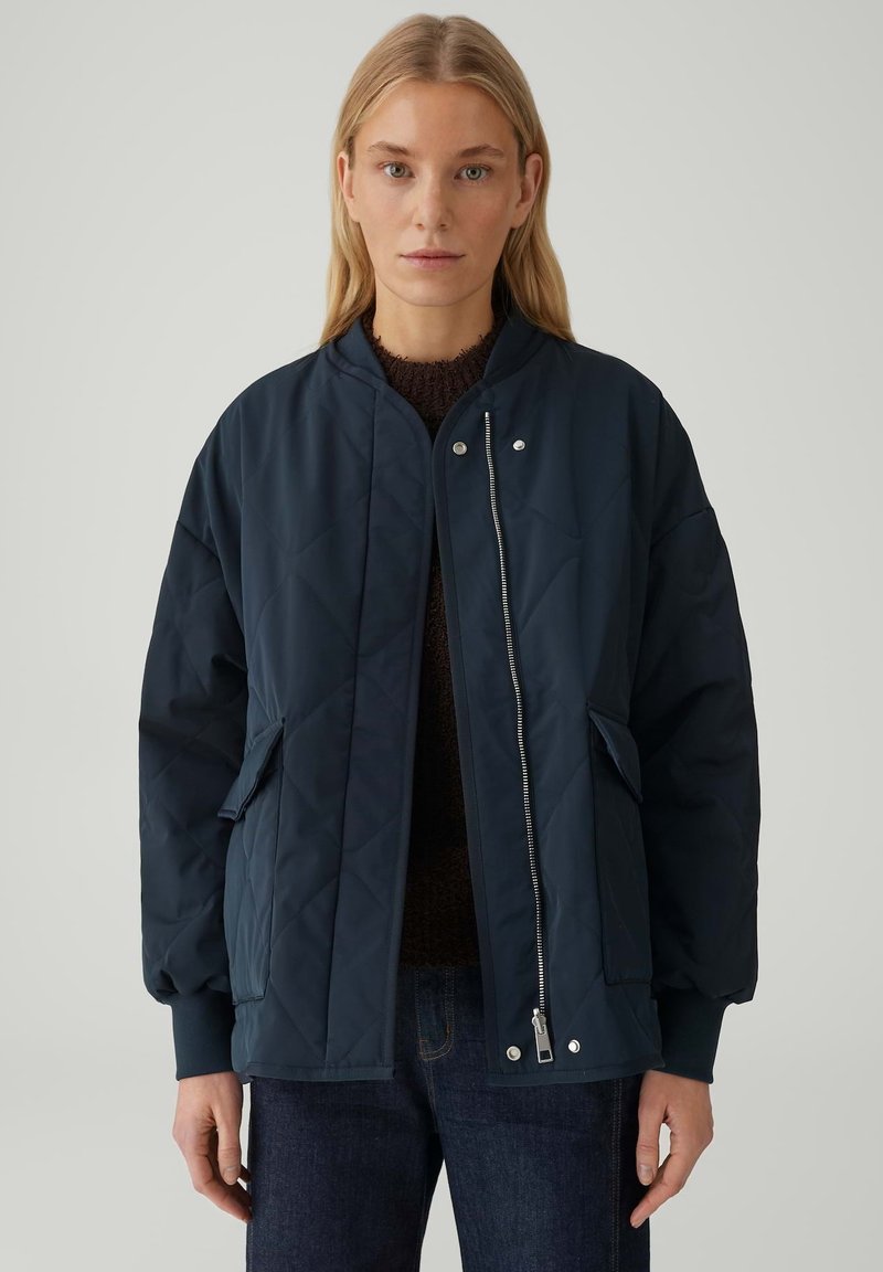 Frau trägt eine marineblaue, gesteppte Bomberjacke mit Fronttaschen, Reißverschluss und Druckknöpfen über einem dunklen Pullover und blauen Jeans.