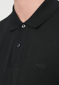 Μαύρο πόλο πουκάμισο από υφασμάτινη υφή με γιακά, διαθέτει δύο κουμπιά και ένα διακριτικό ανάγλυφο λογότυπο στο αριστερό στήθος.