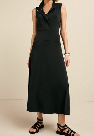 Robe midi noire sans manches avec col et détails de boutons, portée avec des sandales plates noires à brides et un bracelet chaîne.