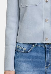 Světle modrá semišová bunda s cropped designem, která obsahuje stříbrné knoflíky a kapsy, kombinovaná s džínami s mírným vyblednutím.