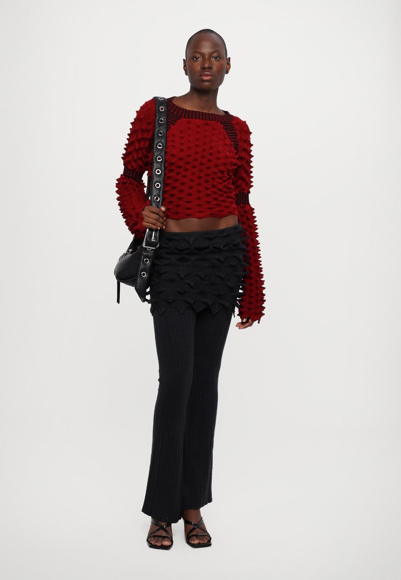 Roter strukturierter Crop-Pullover mit geripptem Kragen, kombiniert mit einem schwarzen geschichteten Minirock und gerippten, ausgestellten Hosen. Schwarze Handtasche und Riemchen-High Heels.