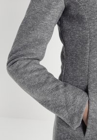 Mouw en zijnaad van een getailleerde grijze textuurjacket met een hand die gedeeltelijk in de zijzak is gestoken tegen een effen achtergrond.