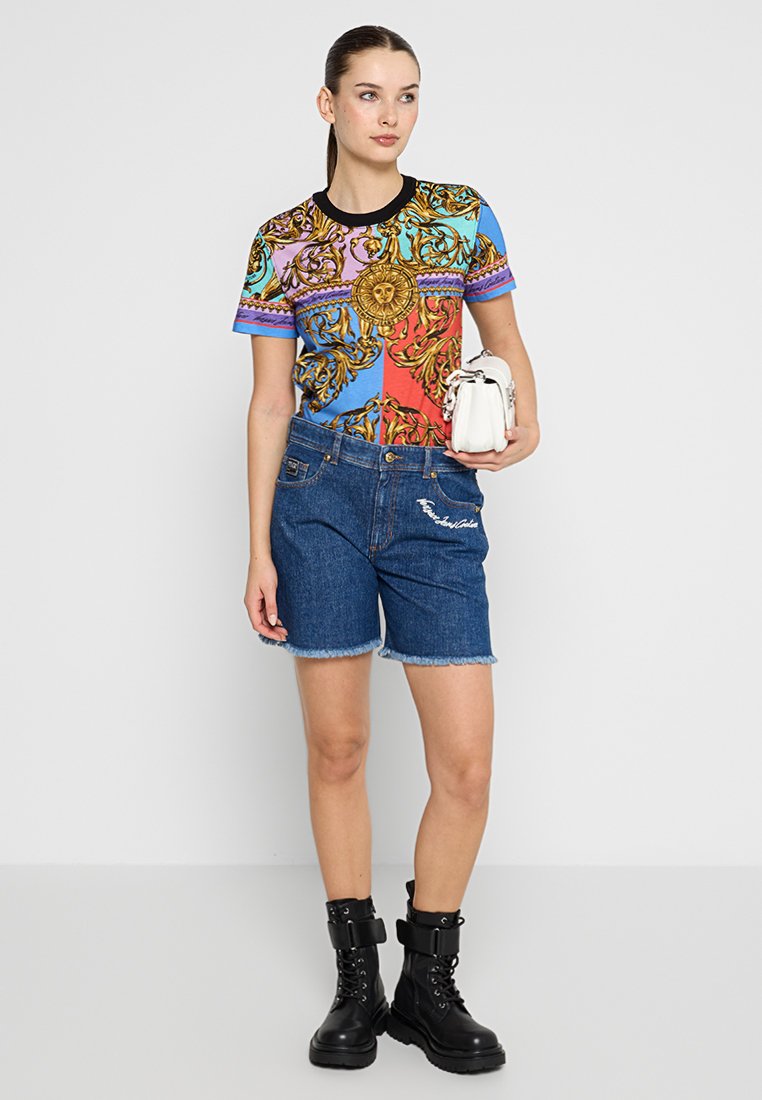 Koszulka o krótkim rękawie w wielokolorową grafikę z ozdobnymi wzorami, zestawiona z postrzępionymi szortami dżinsowymi i czarnymi botkami do kostki. Trzyma małą białą torebkę.