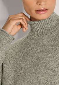 Grauer Rollkragenpullover mit gerippter Textur und leichtem Schimmer, mit enganliegenden Bündchen und lässigem Schnitt. Die Hand des Models ist in der Nähe des Kragens.