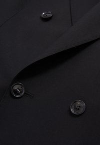 Zwarte dubbele blazer van soepel stof, met klepjes in de revers en glanzende zwarte knopen. Fijne stiksels aan de randen.