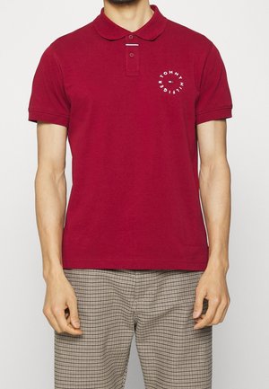 Mężczyzna w czerwonej koszulce polo z krótkim rękawem z okrągłym logo Tommy Hilfiger oraz beżowych, kraciastych spodniach, stojący na białym tle.