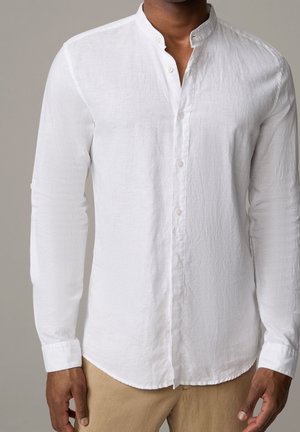 Uomo che indossa una camicia di lino bianca a maniche lunghe con collo alla coreana e pantaloni beige su sfondo neutro.