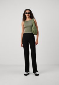 Zelené přiléhavé tílko, černé široké kalhoty s žebrovanou texturou a stříbrné boty na platformě. Zelená texturovaná kabelka přes rameno, zakřivené sluneční brýle, přirozené kudrliny.