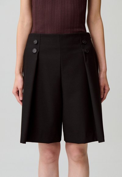 Pantalones cortos negros a medida con un diseño estructurado, plisados en la parte delantera y dos botones decorativos a cada lado. La tela tiene una textura suave.