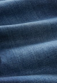 Denim tkanina, tamnoplave boje s glatkom teksturom i blagim sjajem. Vidljiv uzorak tkanja s zakrivljenim nabora i suptilnim varijacijama na površini.