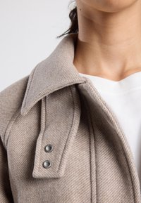 Close-up of beige wool coat collar with metal snap buttons over a white crew neck top on a person’s neck and shoulder.