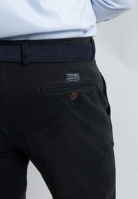 Zwarte broek van geweven stof, met een bruine knoop op de achterzak en een label dat op de tailleband is genaaid, met een gladde textuur.