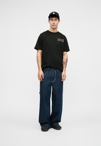 Sort bomulds T-shirt med grafik på brystet, parret med brede denimbukser med striber. Modellen bærer sort kasket og slip-on sko.