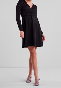 Robe noire en portefeuille avec manches longues, tissu texturé, décolleté en V et jupe fluide. Associée à des sandales à lanières bleues.