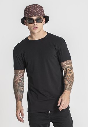 Camiseta negra de manga corta con acentos de patrón de marca en las mangas. Combinada con un sombrero tipo bucket marrón de patrón y gafas de sol.